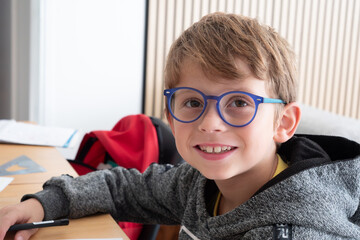 Boy wearing glasses is smiling. He is sitting at a table with a pen and a book