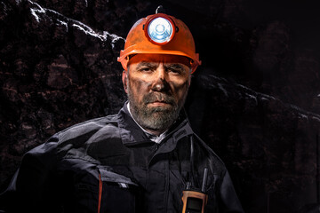 Coal miner with a dirty face in black dust, wearing a protective helmet with a flashlight and work...