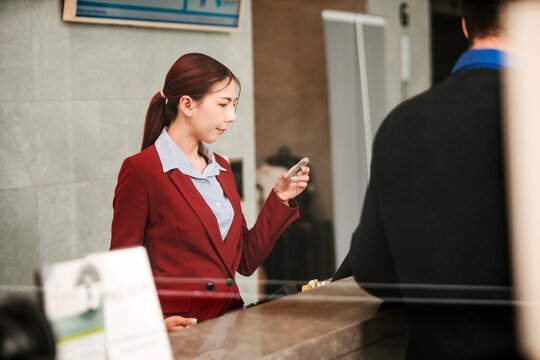 A woman at a hotel reception desk checks her phone while assisting a guest in a business setting, illustrating modern hospitality, service efficiency, and professional front desk interaction.