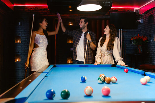 Three friends cheer around a pool table in a stylish, dimly lit billiards lounge, enjoying a night of laughter, competition, and connection in a vibrant nightlife setting.