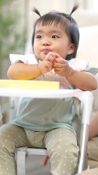 Asian toddler girl sucking thumb while eating fruit, showing natural curiosity and early coordination, self learning moment, healthy development and family lifestyle Vertical