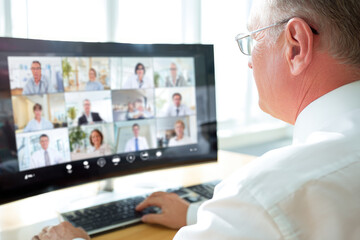 Senior businessman participates in video call meeting with colleagues on computer screen in bright office environment, showing teamwork and communication