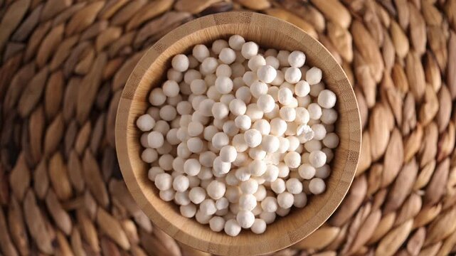 White dry tapioca pearls in wooden bowl rotating on wooden background. Top view of raw tapioca starch balls used for bubble tea or desserts. Tapioca pearls as healthy gluten free ingredient.
