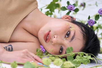 Overhead shot of a young woman touching her face, lying among green plants and purple flowers."