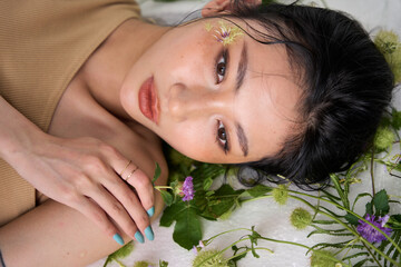 A young woman lying on her side amidst green foliage, gazing intently at the camera with a serene expression."