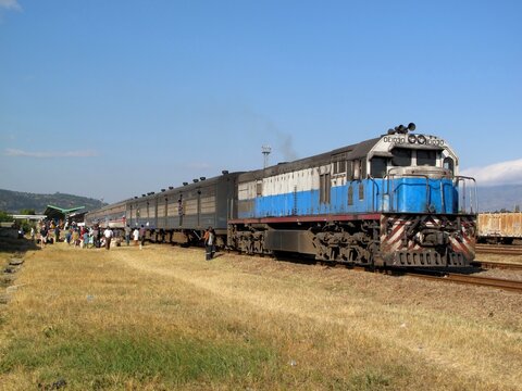 TAZARA train at Mbeya Station in Tanzania, with passengers boarding against a backdrop of hills and blue sky.