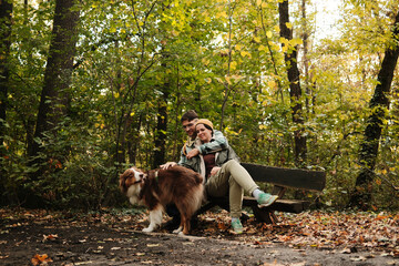 Couple sitting together on wooden bench with Australian Shepherd dog in colorful forest. Concept of love, peace, and connection