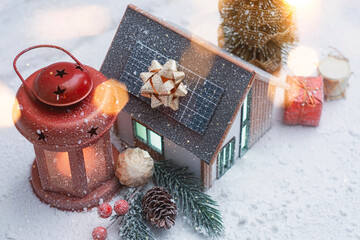 Miniature Christmas house with solar panels on the roof, glowing red lantern and snowy festive decorations. Cozy winter holiday scene for seasonal designs, New Year and Christmas.