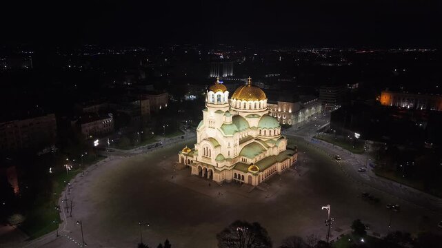 Drone aerial view approaching the illuminated Alexander Nevsky Cathedral from above, showcasing its golden domes and the glowing night cityscape of Sofia.