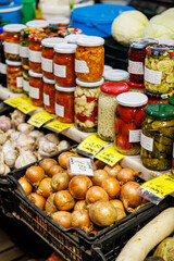 Indoor market stall with crates of apples, onions, potatoes, garlic, root vegetables, herbs, and jars of homemade preserves and pickles