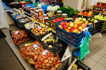 Indoor market stall with crates of apples, onions, potatoes, garlic, root vegetables, herbs, and jars of homemade preserves and pickles