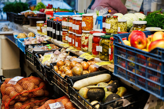 Indoor market stall with crates of apples, onions, potatoes, garlic, root vegetables, herbs, and jars of homemade preserves and pickles