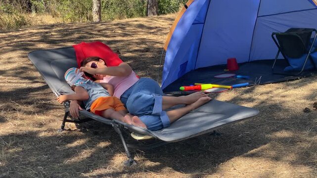 A mother and son lie together on a camping cot. The boy rests on his mom while she relaxes near a tent. They enjoy a quiet moment of bonding in the sunny pine forest.
