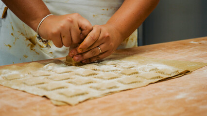 Hands shaping fresh ravioli on a floured wooden board, using a manual cutter to craft rustic, handmade pasta pieces for an authentic Italian kitchen experience