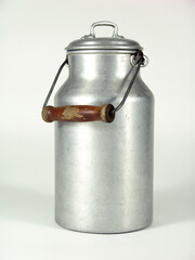 Vintage aluminum milk can with a cylindrical body, rounded lid, and a sturdy metal handle featuring a worn wooden grip, showing signs of age and rustic charm.