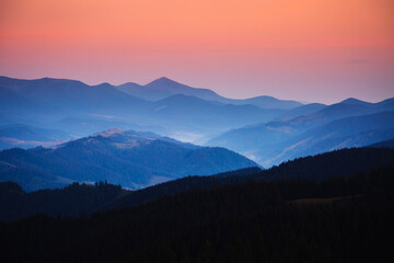 Hoverla peak is visible in the distance among the silhouettes of Carpathian mountain ranges, painted in blue and purple tones in the fading light. Photo wallpaper. Discovery the beauty of earth.