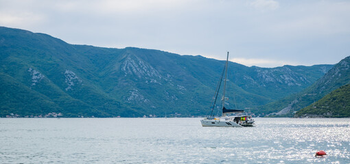 Obraz premium A sailboat glides across the calm waters of Montenegro, surrounded by lush green mountains under a cloudy sky. The serene landscape captures the beauty of the Balkans.