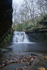Long-exposure of a tranquil woodland waterfall on an autumn day. Moss and leaf covered rocks, soft flowing water, and rich forest textures create a peaceful natural landscape ideal for outdoor themes.