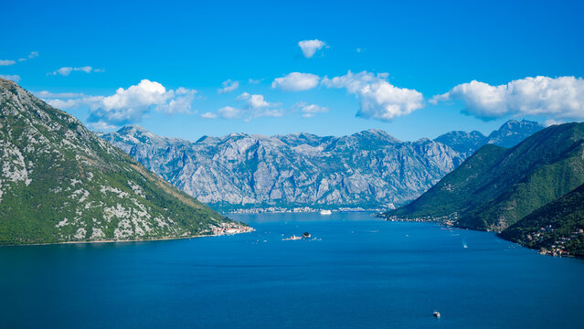 A scenic view of Kotor Bay in Montenegro, surrounded by majestic mountains under a clear blue sky. The calm water reflects the landscape.