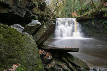 Long-exposure of a tranquil woodland waterfall on an autumn day. Moss and leaf covered rocks, soft flowing water, and rich forest textures create a peaceful natural landscape ideal for outdoor themes.