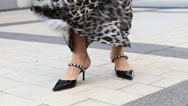 Flowing leopard print maxi skirt moving in wind, woman on city street in elegant high heels black pointed toe shoes. Fashionable detail of stylish and feminine urban outfit