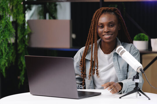 Portrait African female young adult podcaster with laptop and microphone in studio setting, video vlogger