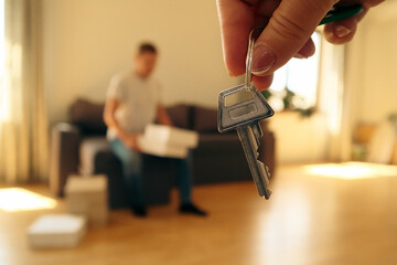 Hand holding apartment keys with person unpacking moving boxes in background.