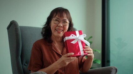 Happy pretty senior asian woman smiling to camera while holding a red gift box with joyful holiday celebration, healthy aging retirement lifestyle gratitude love and festive happiness concept.
