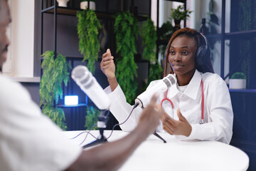 African female doctor in white coat podcast interview with microphones in studio, scientific...