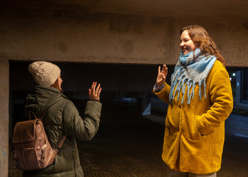 Two women friends saying goodbye and waving to each other at night, wearing warm winter clothes and