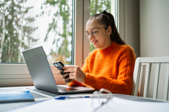 Work from home with phone and laptop. Woman with computer on kitchen table. Entrepreneur making business content with smartphone. Happy freelancer.