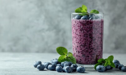 A refreshing blueberry smoothie in a clear glass, garnished with mint leaves and surrounded by fresh blueberries on a gray surface.