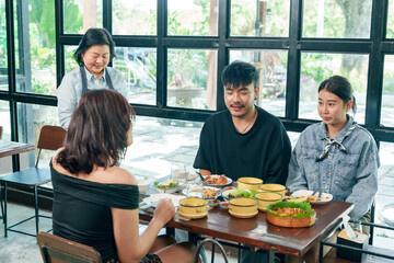Senior asian woman restaurant owner smiling while serving meal to group of adult customers sitting at wooden table inside modern cafe small business showing friendly service authentic food experience