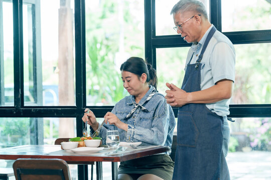 Asian elderly cafe owner engaging with female customer during meal in small restaurant, cheerful moment service, communication between staff diner, collaboration, teamwork, small business management - Powered by Adobe