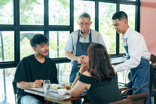 Elderly asian restaurant owner and waiter serving food to couple at table inside cafe teamwork hospitality communication cooperation small business management partnership friendly customer service - Powered by Adobe