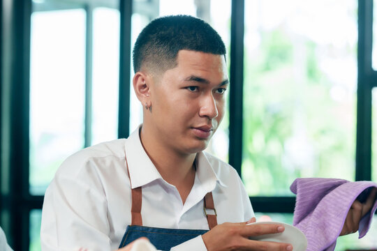 Asian young male barista wearing apron cleaning dish with purple cloth during teamwork session in cafe restaurant small business cooperation family workplace management hospitality service learning