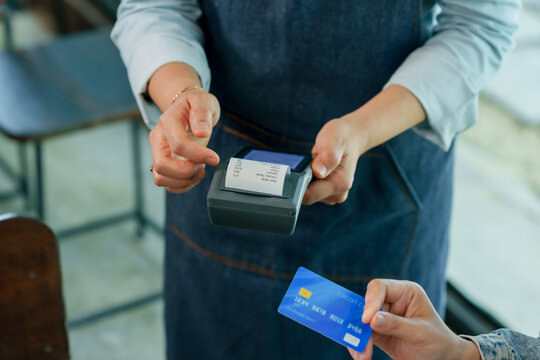 Close up asian woman restaurant owner completing contactless payment with customer using credit card wireless terminal printing receipt representing cashless modern business transaction in cafe