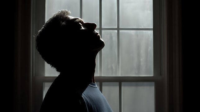 Silhouette of man with head tilted back against window, dramatic lighting creating moody atmosphere for concepts of contemplation, loneliness, or emotional struggle.