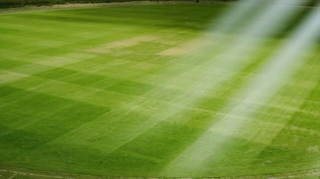 Aerial view of a lush green cricket field with striped patterns in the grass illuminated by soft natural light showcasing vibrant green tones and subtle textures during the day