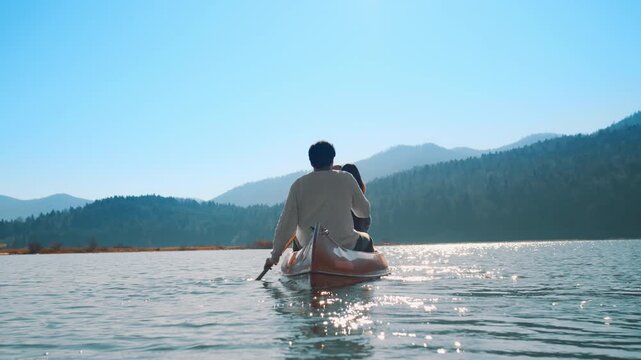 A couple paddles a canoe on a serene lake, surrounded by forested mountains under a clear blue sky. The scene captures a peaceful weekend, emphasizing connection, nature, and an active lifestyle.