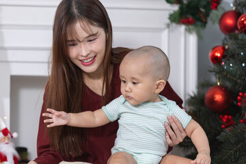 Happy asian young mother holding her baby while playing with Santa toy in cozy Christmas setting surrounded by gifts and decorated tree, capturing joyful holiday family moments at home.