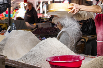 Piles of white flour for sale. A group of women sift white flour behind big piles of it for sale at a stall. A traditional rice vendor serving customers at a local market.
