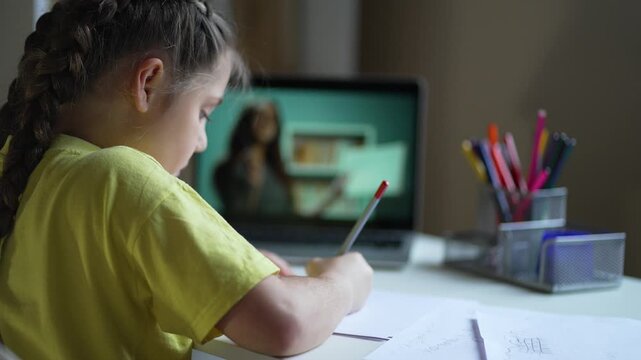 Child writing homework at desk near laptop during online class, girl focused on study and school assignment, distance learning setup shows teacher on screen with pencil paper holder visible workspace