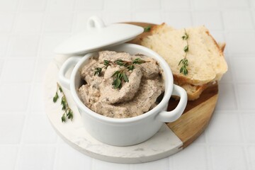 Tasty beef Stroganoff with thyme in casserole and bread on white tiled table, closeup