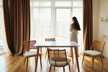 Woman by window in morning interior, standing near dining table and chair with curtains, wearing robe and looking outside at sunlight on wooden floor, peaceful home scene of quiet solitude.