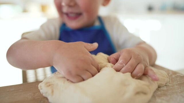 Toddler kneading dough at kitchen table child with blue apron uses hands to press and play with baking dough while smiling in playful activity showing texture and motor skill practice child smile