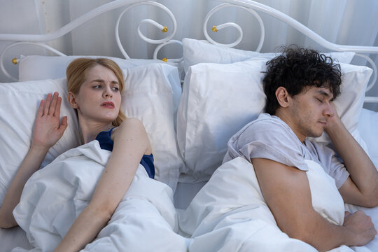 Annoyed women turning away from sleeping man in bed, expressing discomfort and relationship tension. Concept of sleep incompatibility, emotional distance and couple communication issues.