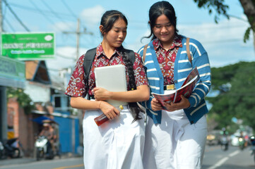 Two students in patterned uniforms, one with a striped sweater, walk along a busy street carrying books and notebooks, discussing lessons on their way home after class.