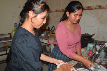Two casually dressed students wash dishes together at the kitchen sink; one hands a plate while the other rinses it under running water, showing teamwork in chores.