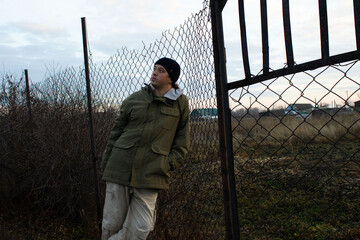 Man in Olive-Green Jacket and Beanie Leaning Against a Weathered Wire Mesh Fence
